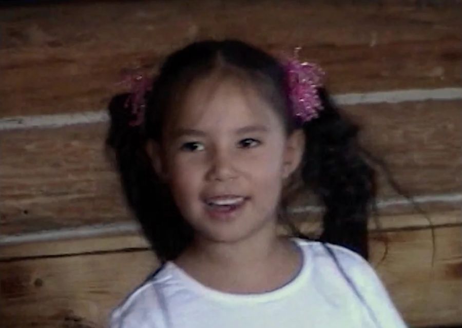 A home-video frame of a smiling young girl in pigtails against a log wall