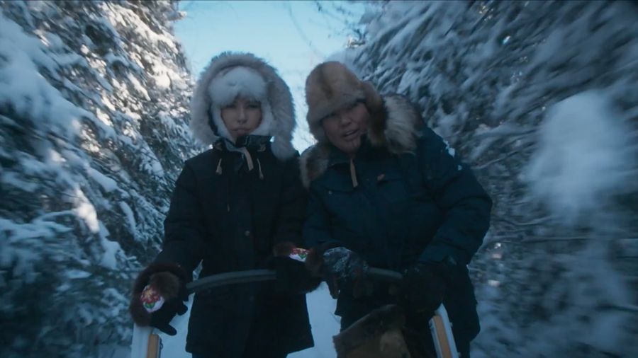 Two women in fur hats on a sled trail through snow-laden spruce